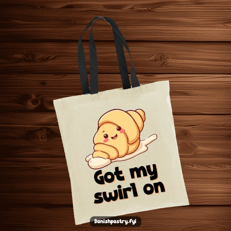 Funny Tote Bag showcasing a smiling croissant character in a playful slide down a streak of icing. A charming and humorous accessory.